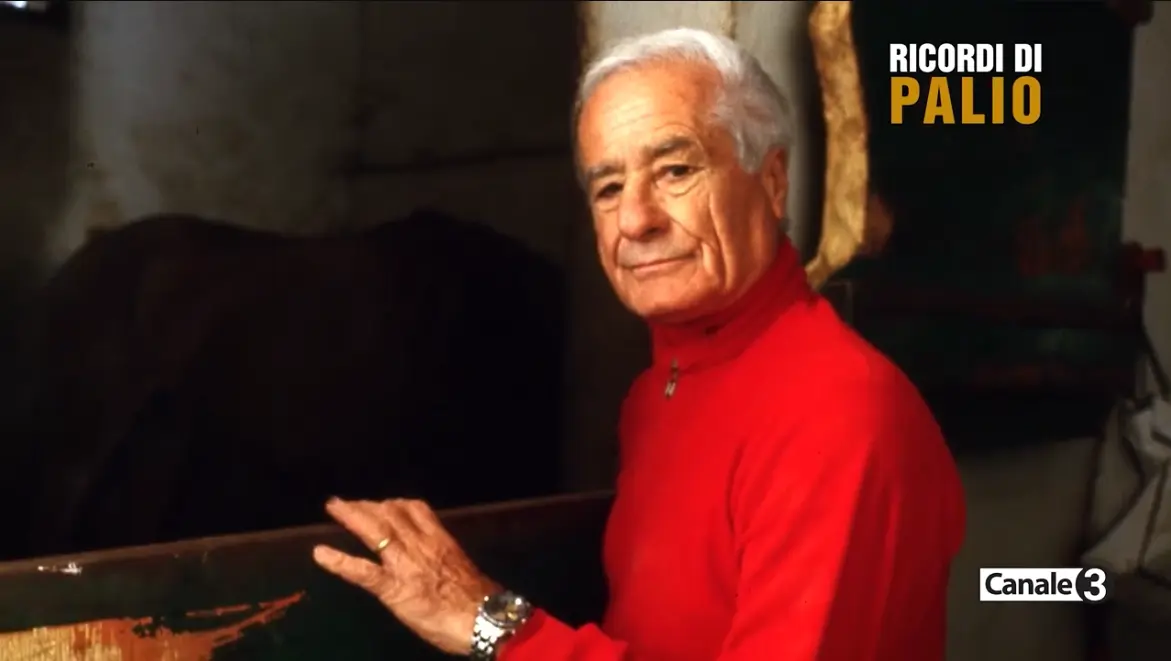 Lazzaro Beligni, detto Giove, in etร matura, ritratto in una scuderia con maglione rosso mentre posa accanto al box di un cavallo, in un fotogramma dellโarchivio Ricordi di Palio.